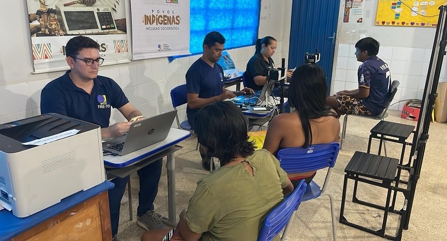 A imagem mostra uma sala para atendimentos. Três servidores, uniformizados e equipados com compu...