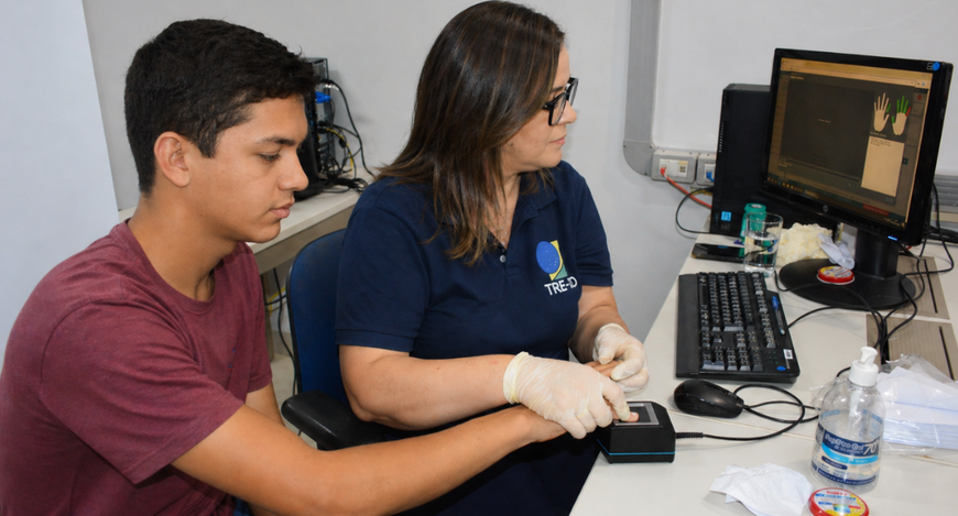 Em um cartório eleitoral, um jovem de camiseta vinho está sentado ao lado de uma servidora que u...