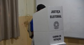 A imagem mostra um aluno votando em uma cabine de votação da Justiça Eleitoral. A cabine é feita...