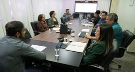 A imagem mostra uma reunião realizada em uma sala de conferências. Nove pessoas estão sentadas a...