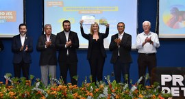 Na imagem, um grupo de seis pessoas posa em um palco durante uma cerimônia de premiação. Ao cent...
