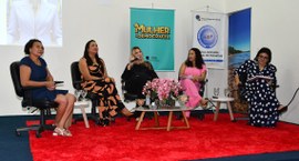 A imagem mostra um evento em ambiente fechado, com cinco mulheres sentadas em cadeiras sobre um ...