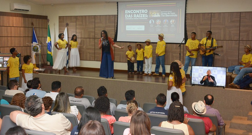 A imagem é uma foto de um evento em um auditório fechado. No centro, há um palco com fundo de pa...
