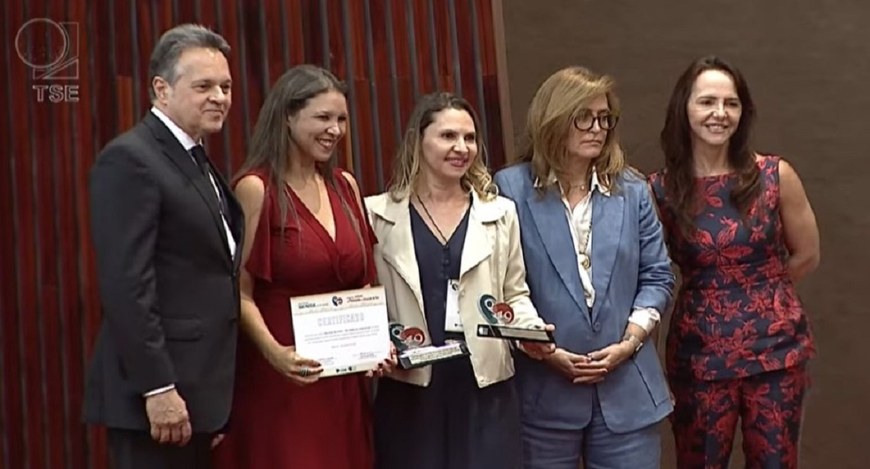 A imagem mostra cinco pessoas posando para uma foto em um ambiente formal. À esquerda, há um hom...