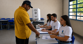 TRE-TO incentiva participação de mesários voluntários nas Eleições 2026 Um homem de camiseta amarela entrega um documento a uma mesária em uma seção eleitoral montada d...