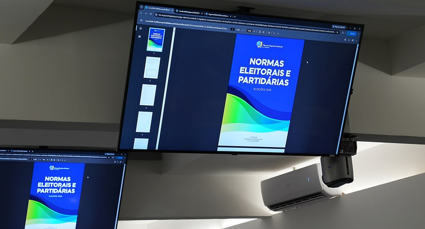Foto da sala de reuniões. Em destaque, um grande monitor exibe a capa da cartilha Normas Eleitor...