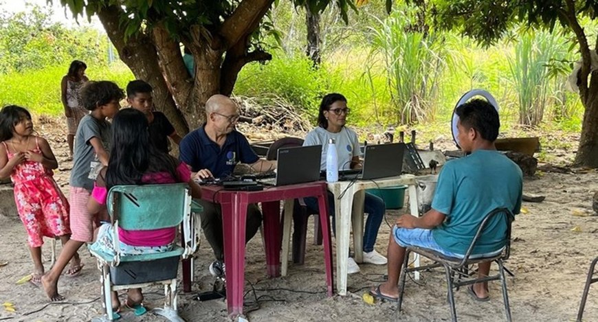 A imagem mostra uma ação de atendimento realizada ao ar livre, sob a sombra de uma árvore, em um...