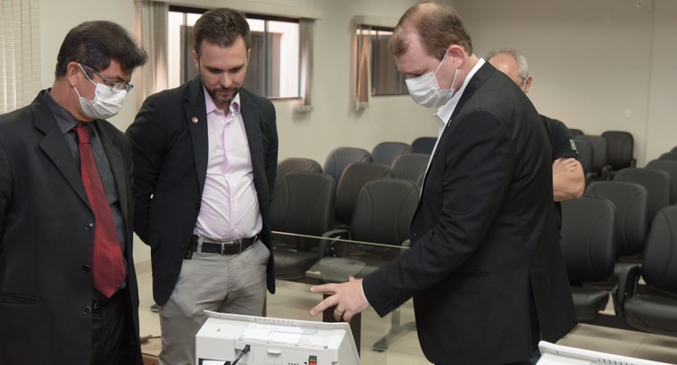 Foto colorida com quatro homens usando ternos ao redor da urna eletrônica.