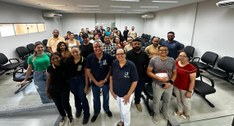 A imagem mostra um grupo diverso de aproximadamente 25 a 30 pessoas reunidas dentro de um auditó...