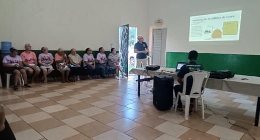 A imagem mostra uma palestra realizada em um salão amplo e simples, com paredes brancas e faixa ...