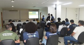 A imagem mostra uma sala com várias pessoas sentadas em cadeiras organizadas em fileiras, todas ...