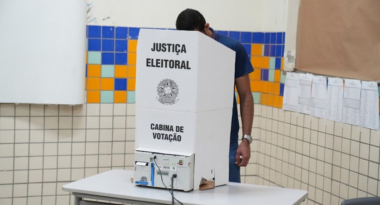 A imagem mostra uma pessoa de pé atrás de uma cabina de votação de papelão branco, colocada sobr...
