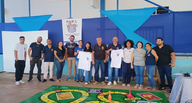 A imagem mostra um grupo de pessoas reunidas em um espaço coberto, uma quadra escolar, posando p...