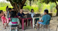 A imagem mostra uma ação de atendimento realizada ao ar livre, sob a sombra de uma árvore, em um...