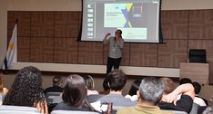 A imagem mostra um auditório durante uma apresentação institucional. Em primeiro plano, aparecem...