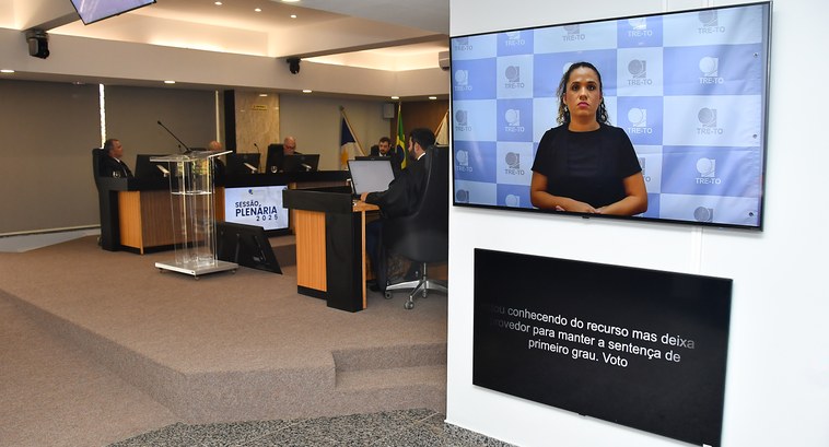 Acessibilidade e Inclusão: TRE-TO faz implantação de painéis de leitura por legenda no edifício sede A imagem mostra uma sessão plenária do TRE-TO, com magistrados em atividade, intérprete de Libra...