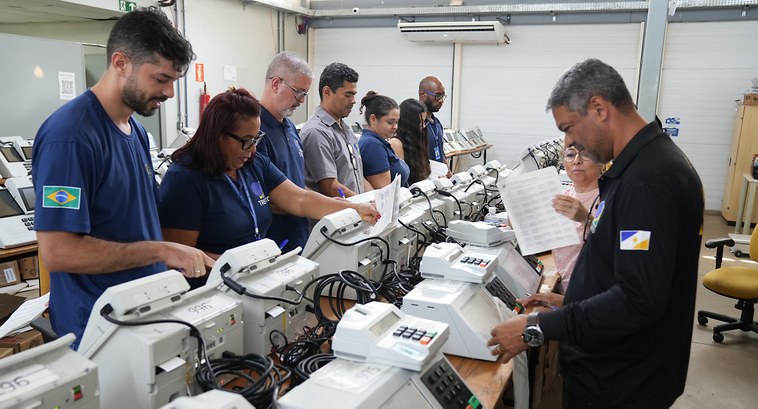 A imagem mostra servidoras e servidores da Justiça Eleitoral realizando testes em urnas eletrôni...