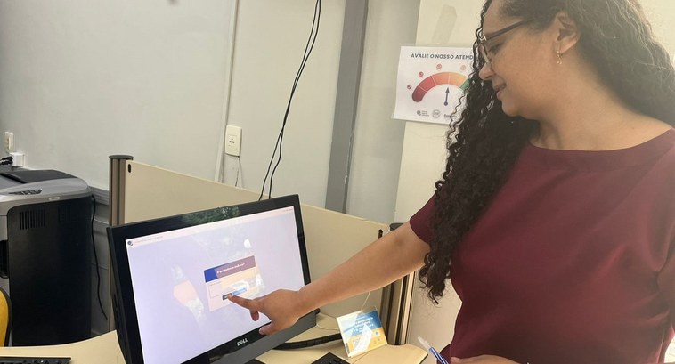 A imagem mostra uma mulher de cabelos cacheados e longos, usando óculos e blusa vinho, interagin...