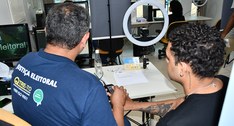 A imagem mostra um ambiente de atendimento da Justiça Eleitoral. Em primeiro plano, duas pessoas...