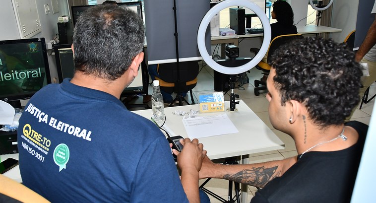 Atenção: prazo para tirar primeiro título e solicitar outros serviços eleitorais vai até 6 de maio A imagem mostra um ambiente de atendimento da Justiça Eleitoral. Em primeiro plano, duas pessoas...