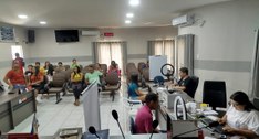 A imagem mostra uma sala de atendimento público. No fundo, há várias pessoas sentadas em cadeira...