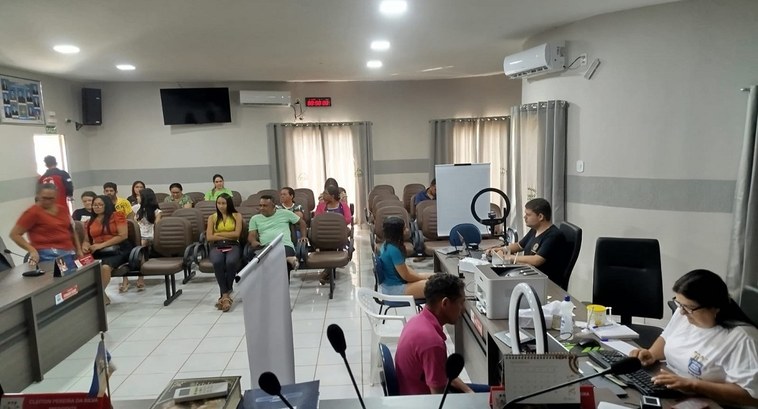 A imagem mostra uma sala de atendimento público. No fundo, há várias pessoas sentadas em cadeira...
