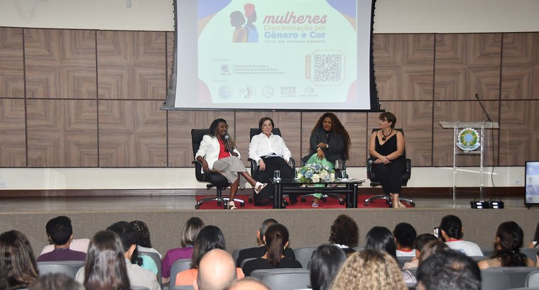 Bate-papo no TRE-TO debate discriminação por gênero e cor enfrentada por mulheres e a importância da educação no combate ao racismo A imagem mostra um auditório com diversas pessoas assistindo a um painel de debate. No palco, qu...