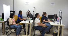 A imagem mostra um ambiente interno de atendimento, organizado em formato de guichês. Quatro mul...