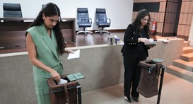 A imagem apresenta duas mulheres no auditório do TRE-TO, colocando cédulas em urnas de lona. Em ...