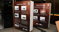 A imagem mostra dois grandes painéis expositivos posicionados em um ambiente interno. Os painéis...