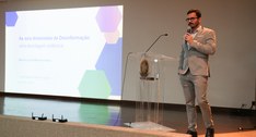 A imagem mostra um palestrante em pé sobre o palco de um auditório, durante uma apresentação rea...