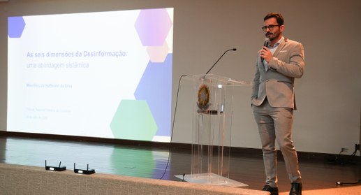 A imagem mostra um palestrante em pé sobre o palco de um auditório, durante uma apresentação rea...