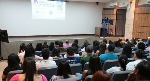 A imagem mostra um auditório durante uma palestra institucional. Em primeiro plano, dezenas de p...