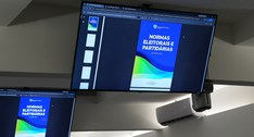 Foto da sala de reuniões. Em destaque, um grande monitor exibe a capa da cartilha Normas Eleitor...