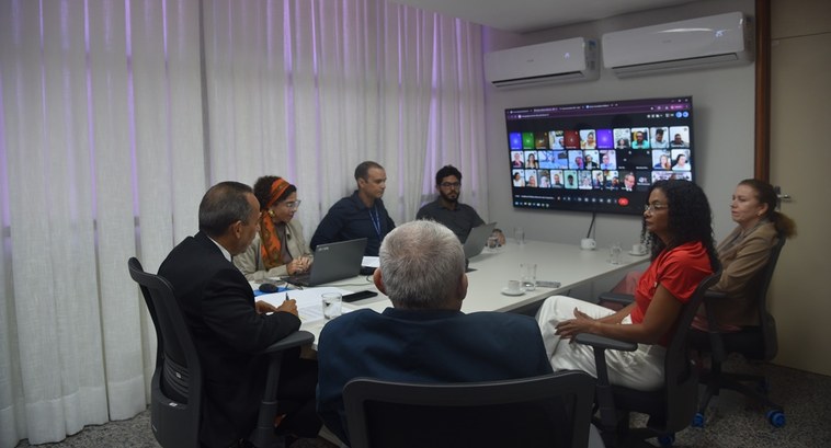A imagem retrata uma reunião institucional em ambiente interno, com pessoas sentadas ao redor de...