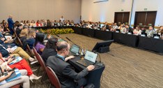 A imagem mostra uma reunião de trabalho realizada em um amplo auditório, com dezenas de particip...