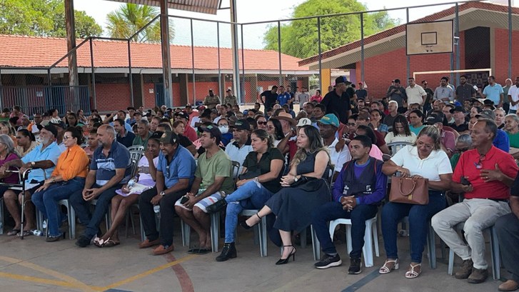 A imagem mostra um grande grupo de pessoas reunidas em um ginásio coberto. Elas estão sentadas e...