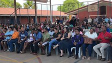 A imagem mostra um grande grupo de pessoas reunidas em um ginásio coberto. Elas estão sentadas e...
