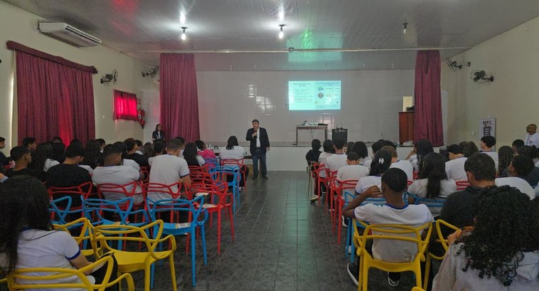 A imagem mostra uma palestra sendo realizada em um auditório escolar. Um homem está em pé à fren...