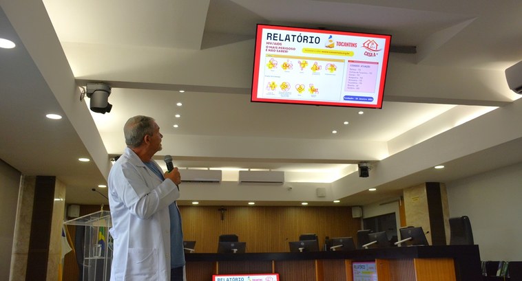 A imagem mostra o médico Alexandre, vestindo um jaleco branco, falando no púlpito do auditório d...