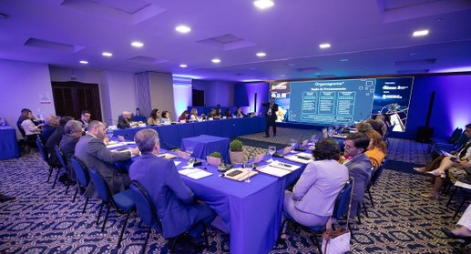 A imagem mostra uma reunião realizada em um salão de conferências iluminado por luzes em tons de...