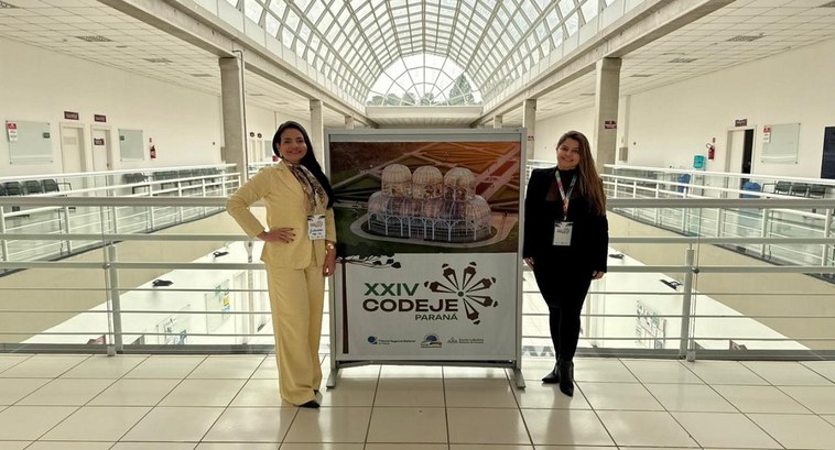 A imagem mostra duas mulheres posando ao lado de um painel do XXIV CODEJE - Paraná, as servidora...