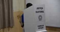 A imagem mostra um aluno votando em uma cabine de votação da Justiça Eleitoral. A cabine é feita...