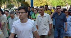 A imagem mostra um grupo de pessoas caminhando em uma área externa arborizada. Em primeiro plano...