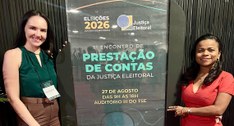 A imagem mostra as duas servidoras posando ao lado de um painel informativo no 1º Encontro de Pr...