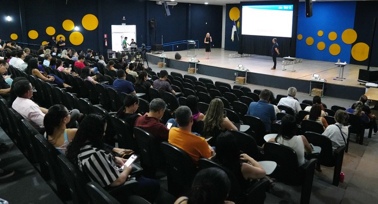 A imagem é um auditório lotado com dezenas de pessoas sentadas em fileiras de poltronas pretas, ...