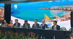 A imagem mostra um painel de debate com sete pessoas sentadas à mesa, em um ambiente formal. Atr...