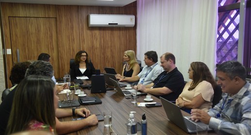 A imagem mostra uma reunião institucional realizada em uma sala de reuniões. Ao redor de uma mes...