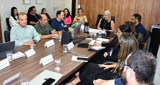 A imagem mostra uma reunião em uma sala de conferências, com um grupo de pessoas sentadas ao red...