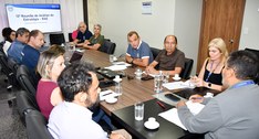 A imagem mostra uma reunião formal acontecendo em uma sala de conferências. Várias pessoas, home...